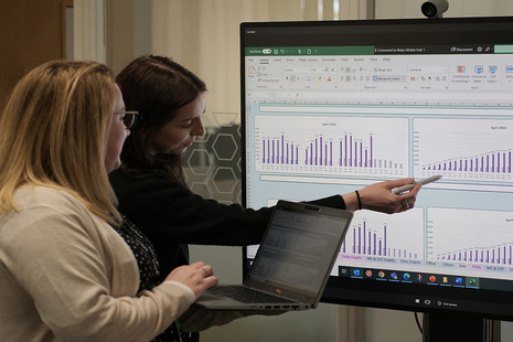Two members of HM Land Registry staff examine figures on a large screen, the woman on the left holding a laptop and the woman on the right pointing at the large screen.